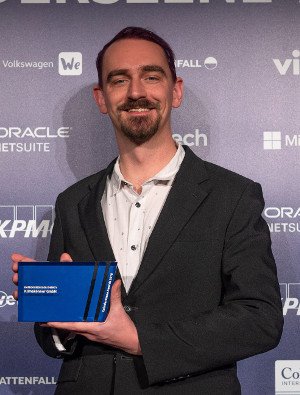 Florian in a suit in front of a blue wall full of sponsor logos, holding a blue plastic award.